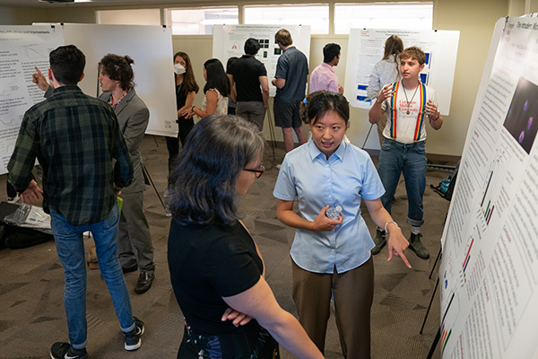 A student points to their research poster while talking to a professor. In the background, pairs of students are gathered around other research posters.