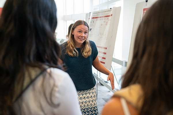 A student facing the camera while talking is framed by two other students facing away from the camera, with only the backs of their heads visible. Two research posters are partially visible in the background.