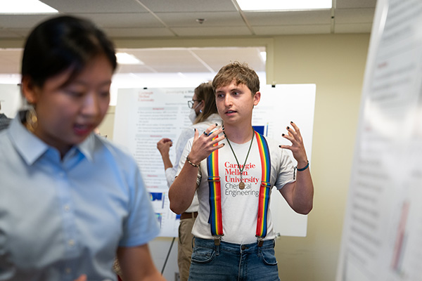 Gabe Mendez-Sanders gestures while standing. Blurry in the foreground are a research poster and a student.