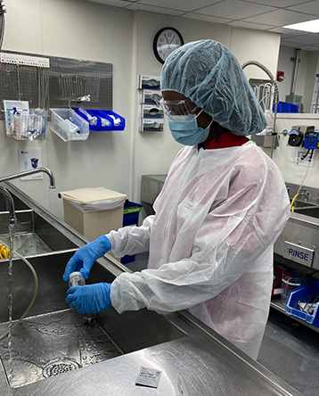 Teresa Myrthil working in a wet lab. She wears a hair net, safety glasses, face mask, lab coat, and gloves.