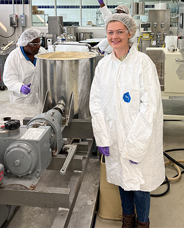 A young woman wearing a white lab coat and hair net stands in an industrial kitchen.