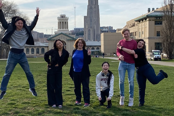 Six adults jumping, hugging, and making silly poses on a lawn outside university buildings.