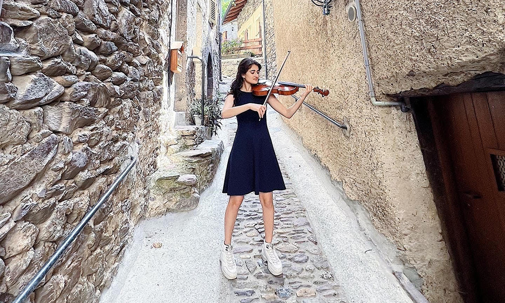 Eleanor David plays the viola standing on a cobblestone sidewalk between two old stone buildings.