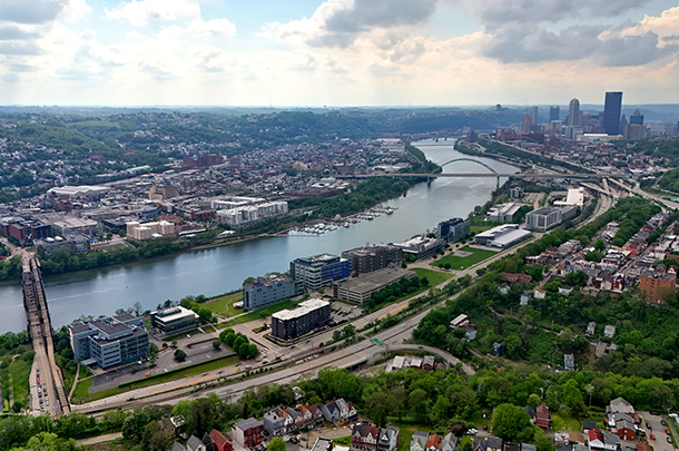 Aerial view of a river, two bridges, city buildings, and bright clouds in a light blue sky.