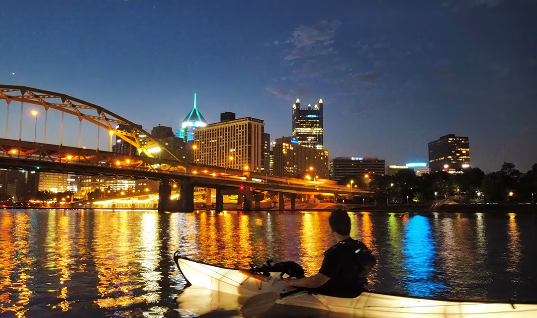 Robert Burnley sitting in an illuminated kayak on a river, with his back toward the camera. In the background, the Pittsburgh skyline is lit up at night.