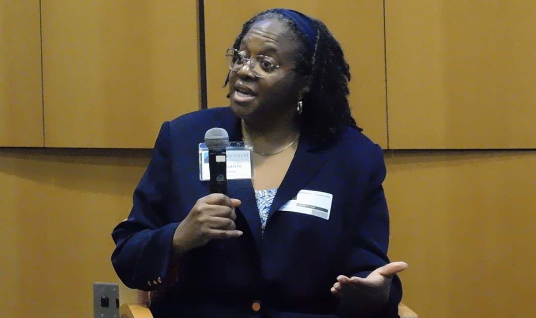  Stephanie Brandford holding a microphone with one hand and gesturing with the other hand while talking.