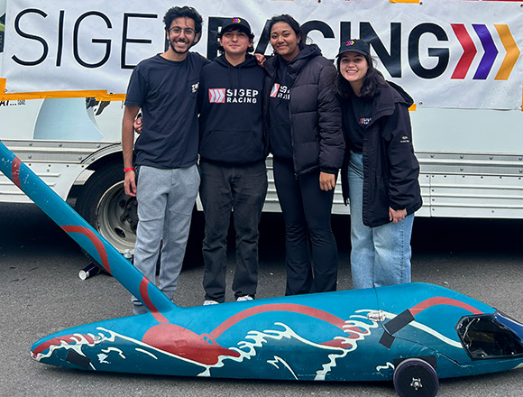 Four college students pose for a photo behind a carbon fiber racing buggy. A banner on the truck behind them reads “Sig Ep Racing.”