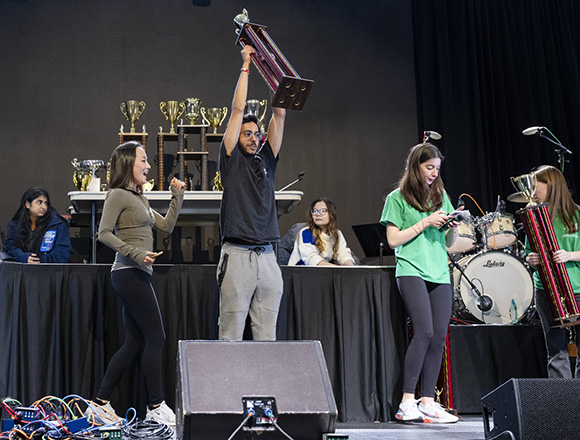 Bilal Chaudhry stands on a black stage with speakers and a drum set, lifting a large trophy above his head.