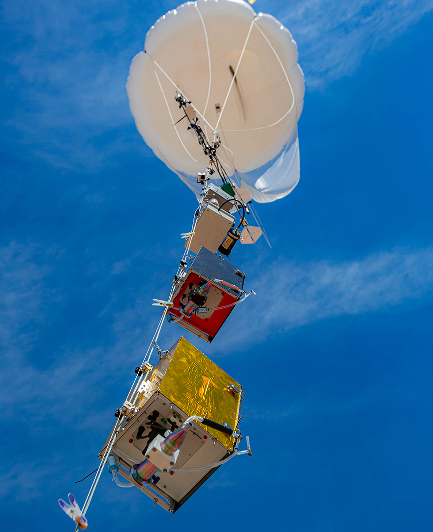 The big white tethered balloon flies in a bright blue sky with multiple scientific instruments hanging from its tether.