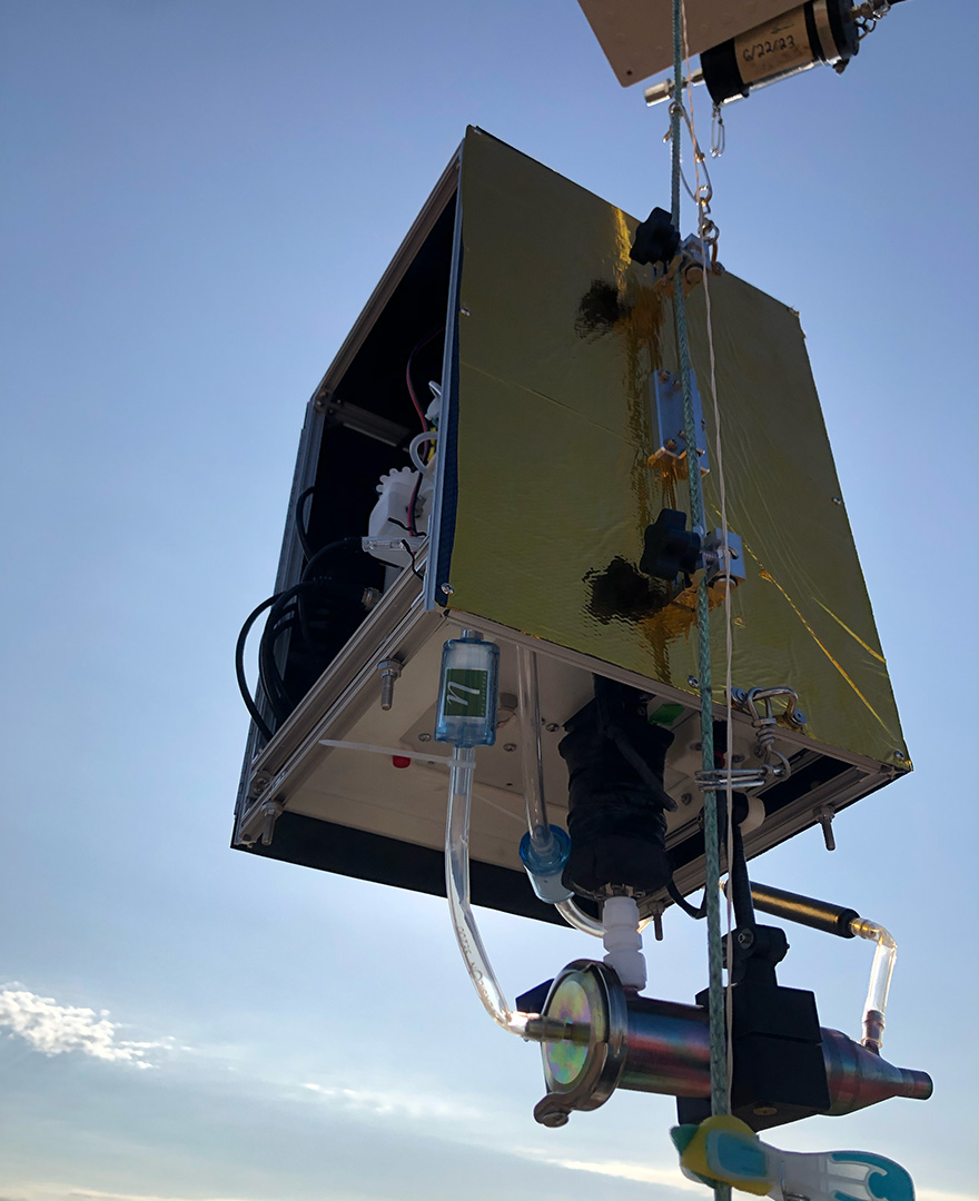 A gold-colored box with small tubes coming out of it, hanging from a tether in the sky.