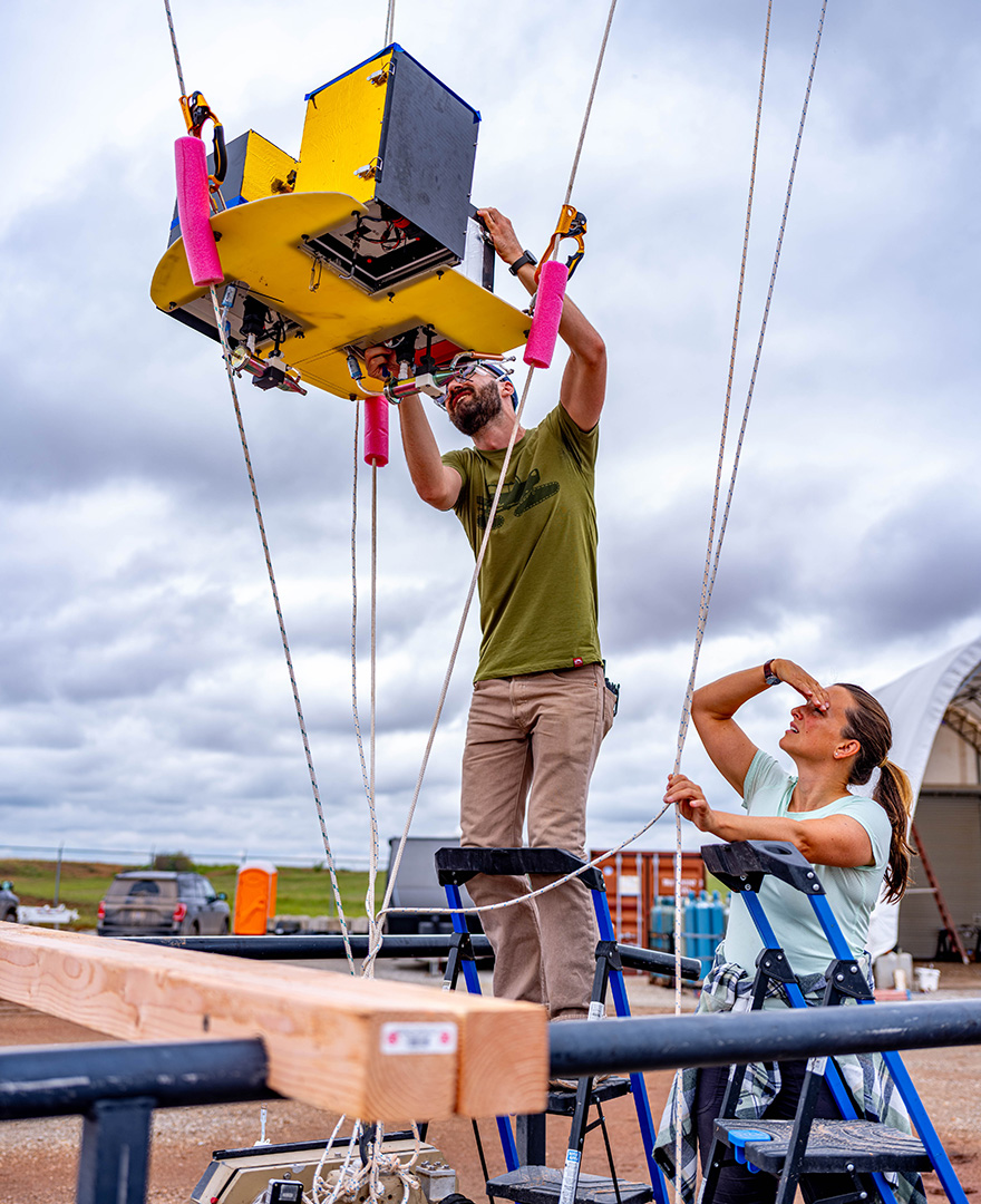 Two adults stand on stepladders to work on a scientific instrument hanging in the air.