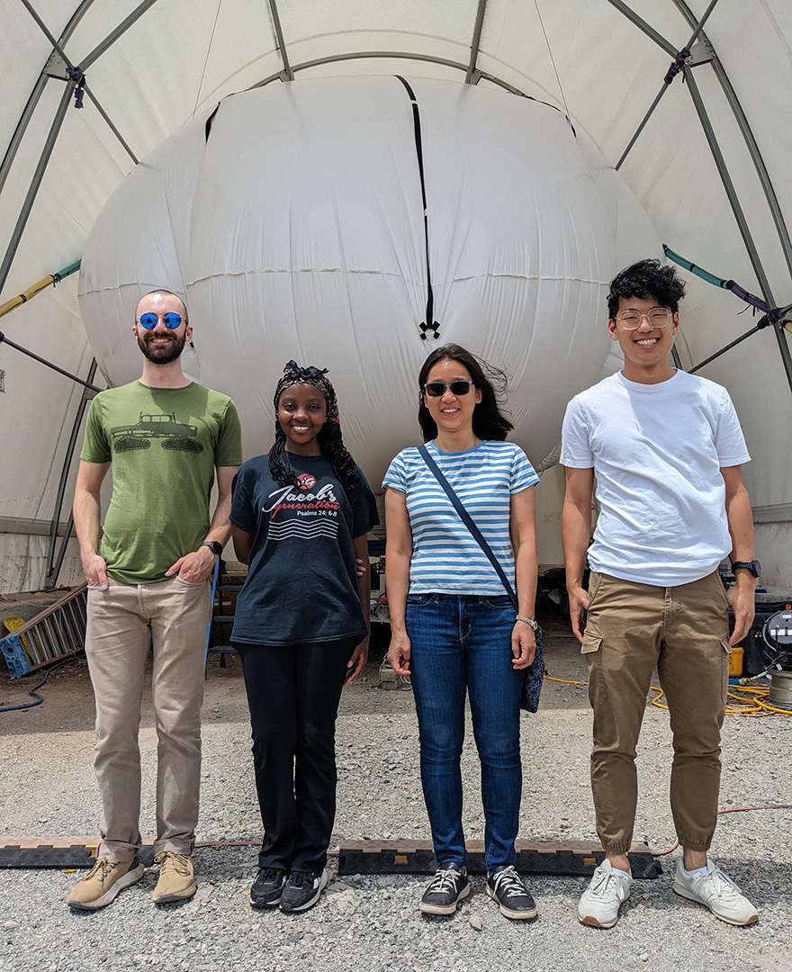 Four people stand smiling in front of a hanger filled by a giant white balloon.