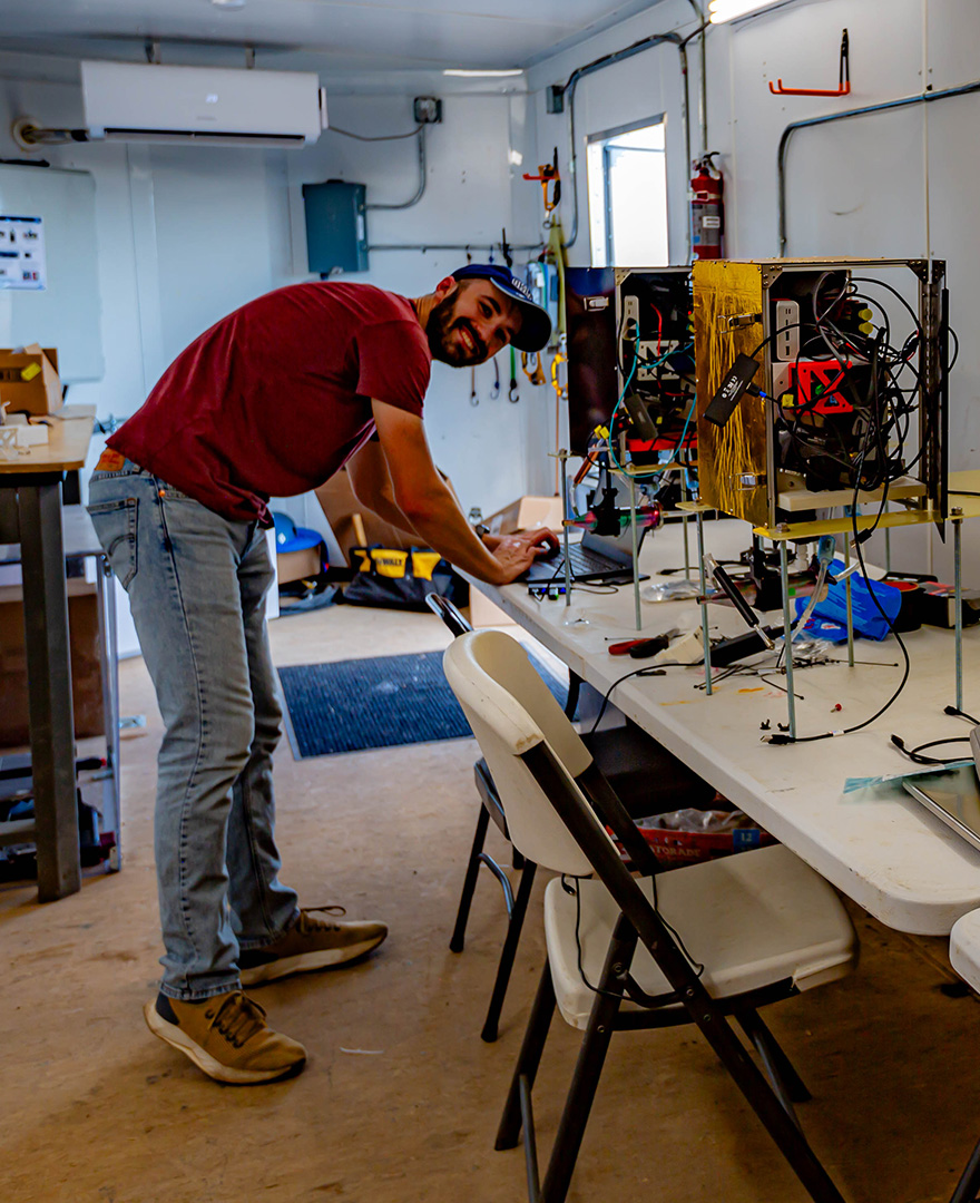 A man smiles at the camera while bent over a table while working on a laptop next to scientific instruments.