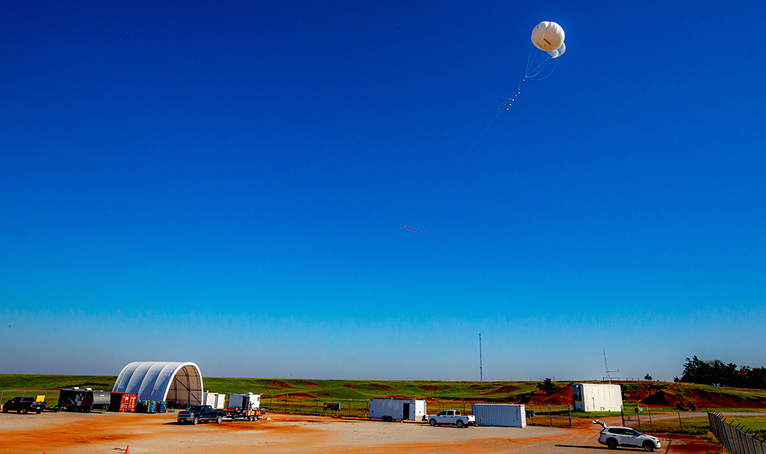 A white tethered balloon flies in a bright blue cloudless sky, above green fields, red clay, and white trailers of equipment