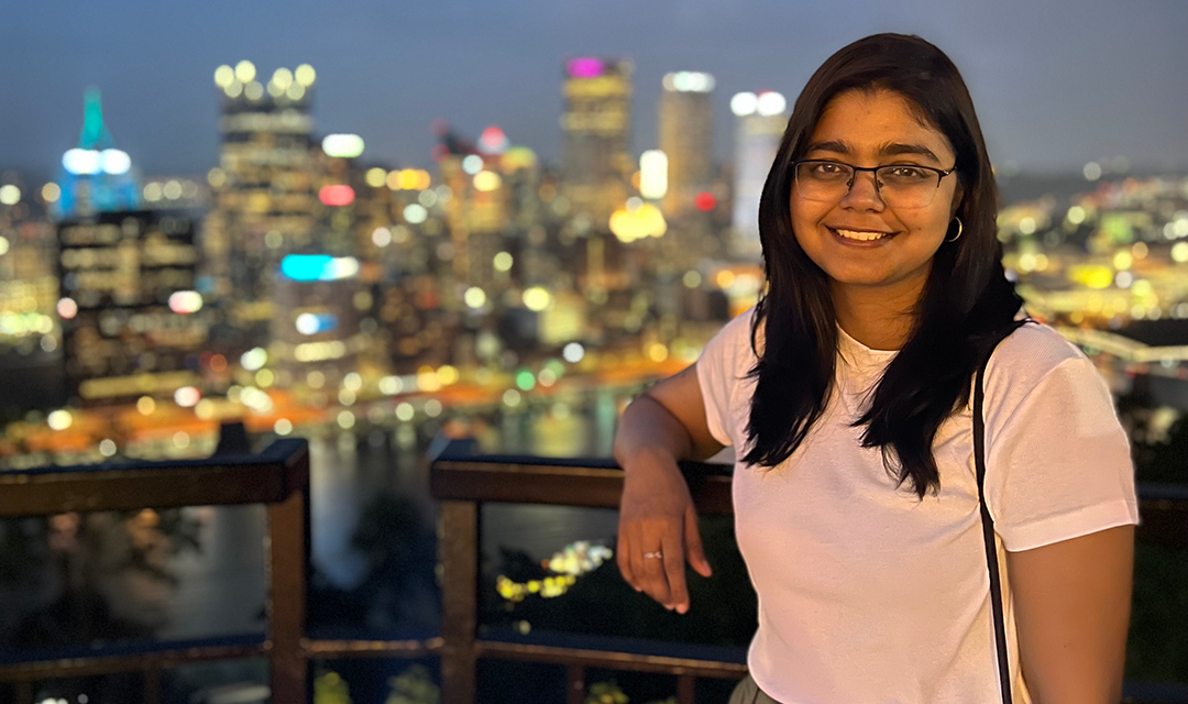 Ashlesha Tiple stands on an overlook with the Pittsburgh skyline behind her, lit up at night.