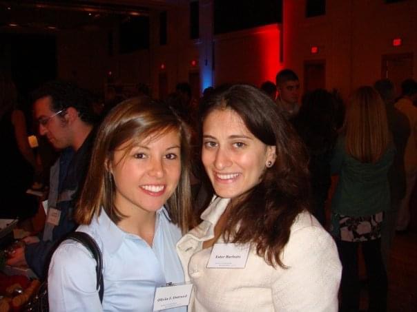 Two young women pose for a photo in a darkened room with other people barely visible in the background.