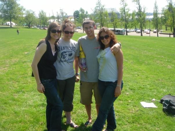 Four college students pose for a photo standing on a grassy lawn, holding a small rocket made with a plastic water bottle and a yellow cone.