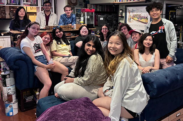 14 college students smile for a group photo while seated on a dark blue couch in a living room with bookshelves and monitors in the background.