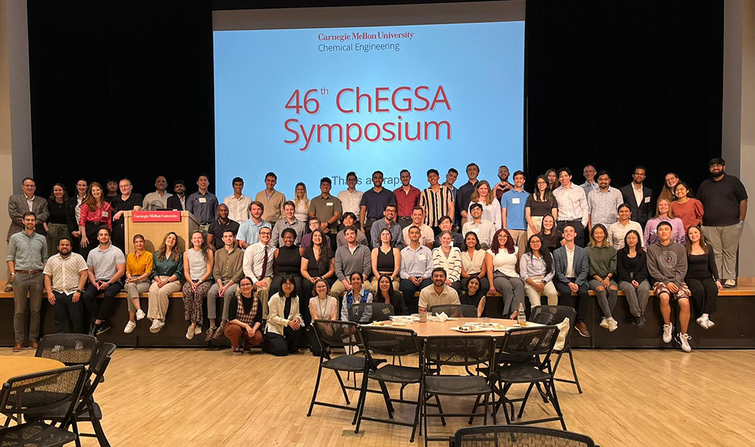 More than 70 students, faculty, and alums seated, kneeling, and standing in rows for a group photo. A screen in the background displays “46th ChEGSA Symposium.”