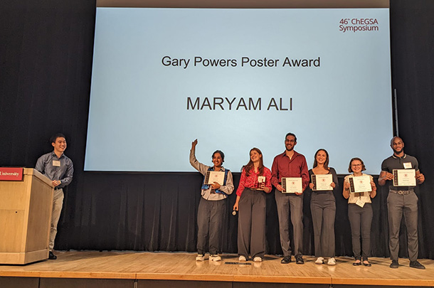 One person stands at a podium, and six people stand in a line on a stage, holding award certificates.