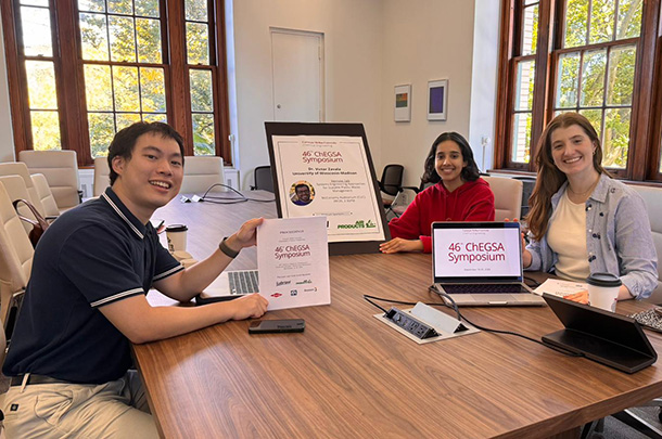 Three people sit at a conference table. On the table are posters and a laptop displaying slides for the ChEGSA Symposium.