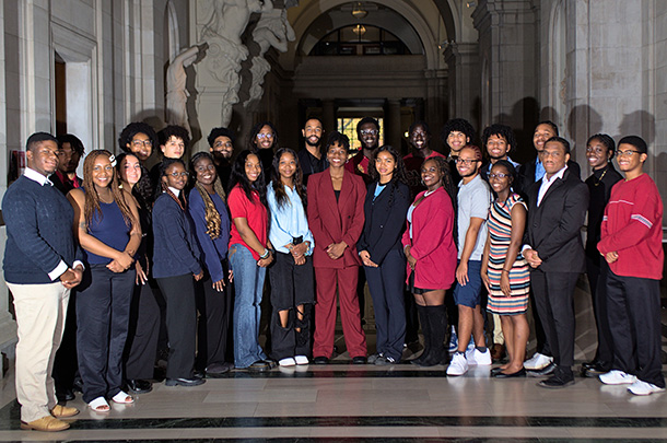 A group of about two dozen college students, some dressed in business attire, pose for a photo in an arched marble hallway.