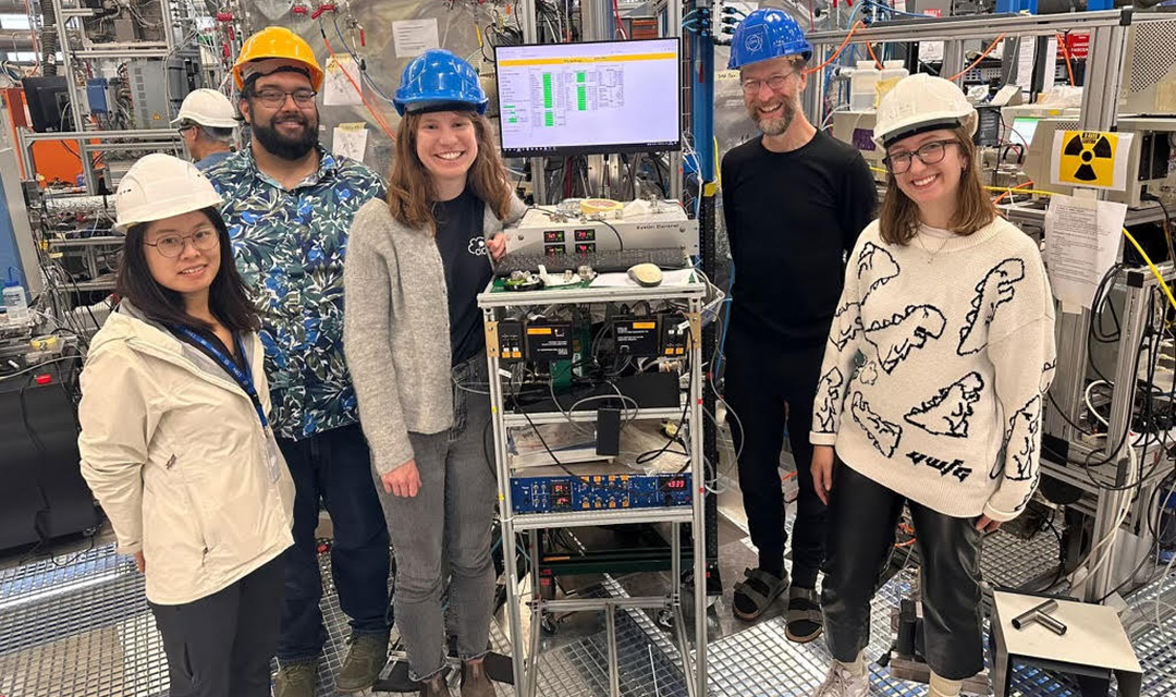 Five researchers wearing hardhats smile for a photo while clustered around a rack of scientific instruments. A computer monitor is mounted on top of the rack, showing a data spreadsheet. The base of a large, silver, cylindrical chamber can be seen behind them, attached to many instruments.