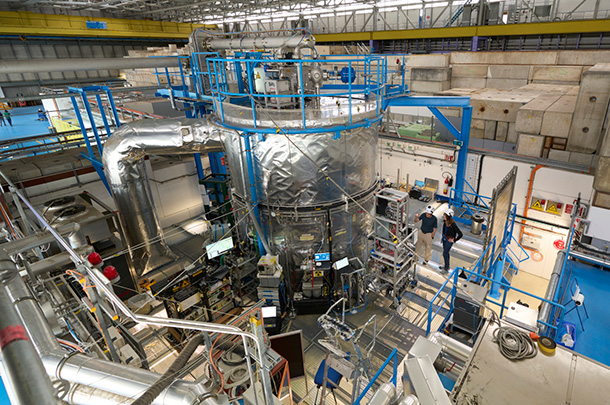 A large, silver cylindrical chamber in a large, warehouse-looking building. Numerous scientific instruments are on racks on the floor around the base of the chamber, attached to it. A large air duct comes off the left side of the chamber and down to the floor.