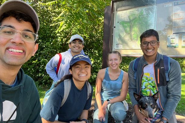 Five college students and one dog pose for a photo in front of a trail map on a sunny day in the woods.