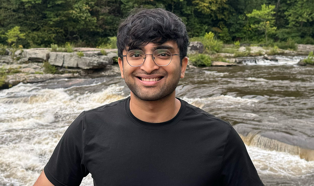 Ali Asger stands on the bank of a shallow river rushing over rocks, with green trees in the background.