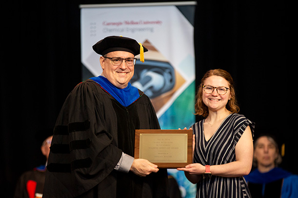 A faculty member wearing regalia hands Megan Walsh an award plaque.