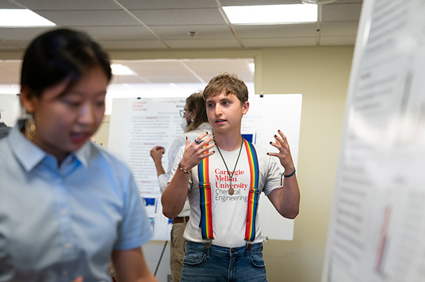 Gabe Mendez-Sanders gestures while standing. Blurry in the foreground are a research poster and a student.