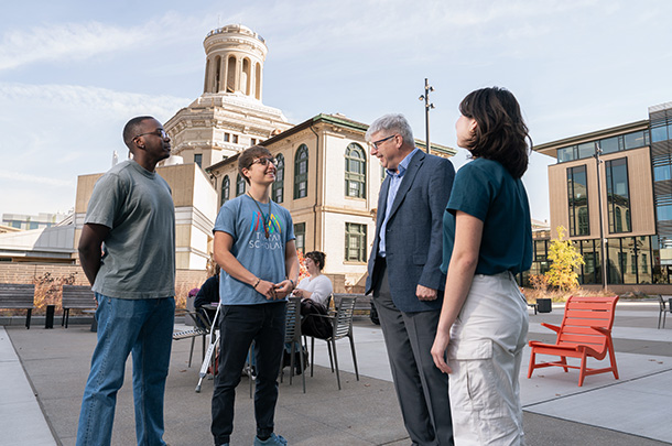 Three college students talk with the dean, standing on a patio outside college buildings