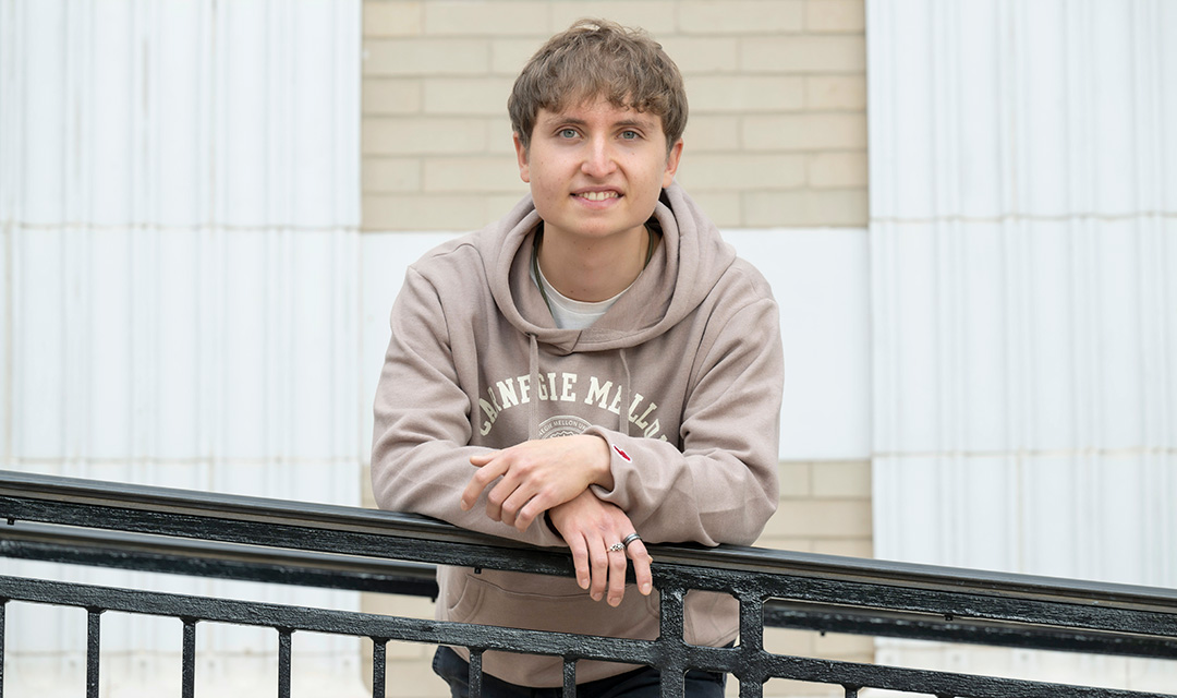 Gabe Mendez-Sanders leans on a black railing in front of a light-colored brick wall