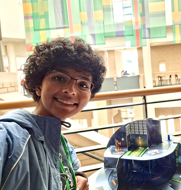 Savi Thompson smiles for the camera and leans on a table on which a Star Wars game is set up.