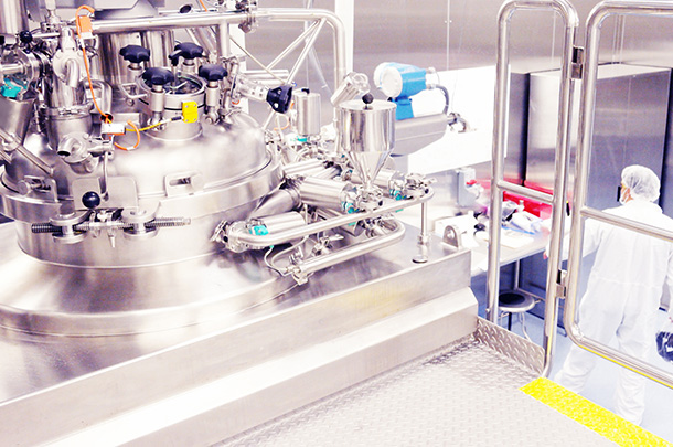 In the foreground of a spotless room is a shiny steel tank with lots of valves and tubes. In the background and off to the right side, a person can be seen from behind, wearing a white lab suite and hair net.